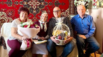 Ювілярка, її родичі та працівник управління соціального захисту усміхаються, позуючи для спільного фото. Пані Катерина тримає в руках пам’ятну монету від міста, її родичка – великий букет квітів, а Іван Фасольник – великий кошик з фруктами.