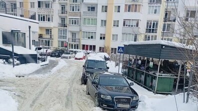 засніжений двір багатоповерхових житлових будинків. Уздовж вузької внутрішньої дороги щільно припарковані приватні легкові автомобілі темного кольору. Машини стоять перед майданчиком для збору відходів.