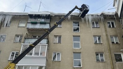 багатоповерховий житловий будинок на даху якого багато нависань буруль. На передньому плані автовишка підняла працівника до рівня даху, щоб збивати бурулі