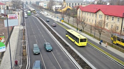 Широка відремонтована вулиця у Львові з кількома смугами руху. Посередині їде жовтий міський автобус, поруч рухаються автомобілі. Праворуч видно трамвайну колію, тротуар із пішоходами та будівлі з червоними дахами. Удалині — будівля головного залізничного вокзалу