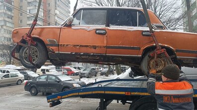 Стара помаранчева легкова машина зі слідами корозії піднята в повітря в процесі евакуації і переміщення на вантажівку. Працівник ЛКП 