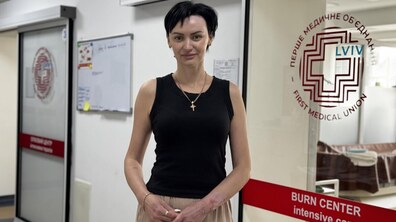 Євгенія Харченко стоїть у холі центру термічної травми та пластичної хірургії лікарні святого Луки. Жінка замислено дивиться в камеру й усміхається, позаду видно лікарняні двері й логотипи Першого медоб’єднання Львова.