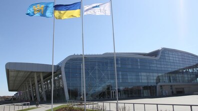 Термінал міжнародного аеропорту Львів. Велика скляна будівля, перед якою стоять три прапори: Львова, України та аеропорту «Львів»