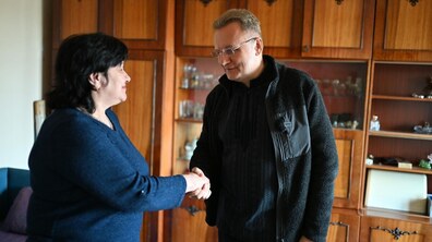 Андрій Садовий тримає за руку Надію Ванчицьку у її новому помешканні. Вони стоять один навпроти одного, обоє усміхаються, що створює відчуття теплої, особистої зустрічі. Андрій Садовий одягнений у темний светр і чорну куртку. Він трохи нахилився вперед під час рукостискання. Надія Ванчицька — жінка з темним коротким волоссям, одягнена в темно-синю кофту. Позаду них велика дерев’яна стінка з шафами та полицями, де стоїть посуд і різні дрібні речі.