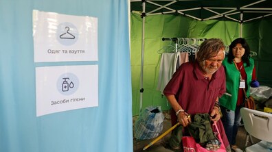 Мобільний простір підтримки з одягом, взуттям та засобами гігієни. Чоловік обирає собі речі, а поруч соціальна працівниця допомагає йому. У наметі на задньому плані – розвішаний одяг.