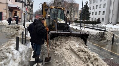 Частина вул. Пекарської. Дорога й тротуари вкриті мокрим, бруднуватим снігом. У центрі – жовтий трактор із великим металевим відвалом спереду. Він рухається вздовж тротуару й згортає важкий мокрий сніг у велику купу.