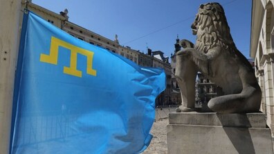 Кримськотатарський прапор майорить на тлі історичної архітектури міста. Поруч із прапором видно кам’яну скульптуру лева біля міської Ратуші.