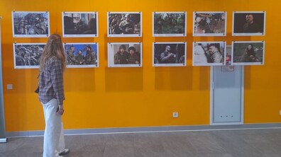 Дівчина розглядає фотовиставку в приміщенні з яскраво-жовтою стіною. На стіні у два ряди розвішані дванадцять світлин у білих рамках, на яких зображені військові