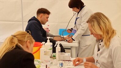 У медичному наметі лікарі обстежують пацієнтів. На передньому плані медична працівниця готує інструменти для забору крові у жінки, яка сидить перед нею. На задньому плані інша медична працівниця вимірює артеріальний тиск молодому чоловікові.