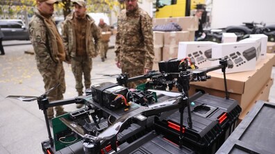 На великих чорних ящиках стоять дрони та інша потрібна військовим техніка. Поруч стоять троє чоловіків у військовій формі.