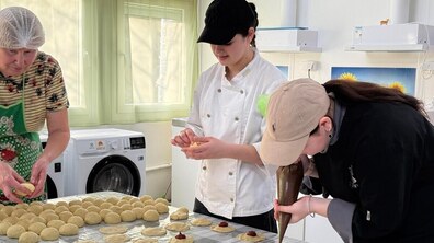 Три жінки спільно працюють за столом: одна формує кульки з тіста, інша розкочує їх, а дівчина за допомогою кондитерського мішка викладає темну джемову начинку. На передньому плані акуратними рядами розкладені заготовки з тіста, готові до подальшого випікання.