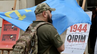 Вшанування пам’яті жертв геноциду кримськотатарського народу біля міської Ратуші у Львові. На передньому плані стоїть чоловік у військовому одязі, який тримає у руках великий блакитний прапор із жовтою кримськотатарською тамгою. В кадрі також два стенди на дерев’яних мольбертах з написами «Геноцид 1944 , Геноцид 2022» у чорно-червоних кольорах, а також «1944 QIRIM» та зображенням колючого дроту.