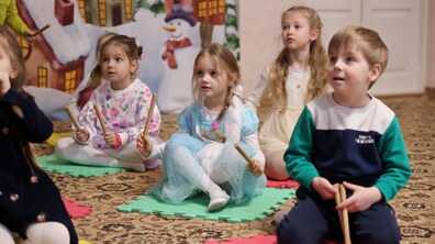 Діти дошкільного віку під час музичного заняття у дитячому садку. Малюки сидять на кольорових килимках із дерев’яними паличками в руках та уважно слухають вихователя.