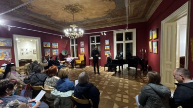 У вишуканій залі з розписаною стелею, роялем та бордовими стінами, прикрашеними картинами, проходить камерний культурний захід. Напівколом затишно розсаджені глядачі, які уважно слухають виступ чоловіка в темному одязі та акомпаніаторки за роялем