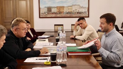 Мер Львова Андрій Садовий, у чорному одязі та окулярах, сидить ліворуч за великим дерев’яним столом і спілкується з мешканцями під час особистого прийому. Навпроти нього чоловік тримає відкриту папку з документами. За столом також присутні інші учасники зустрічі. На столі розкладені документи, папки, блокноти та пляшки з водою.