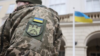 Рука військового з нарукавним знаком Державний Прапор України та нарукавним знаком військової частини на фоні Державного Прапора України на флагштоку діля Львівської міської ради.