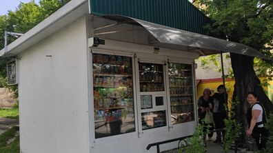 Кіоск, в якому продають продукти через невелике віконце. Біля нього стоять жінки. Поруч росте велике дерево