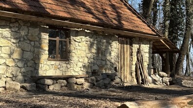 Невеликий будиночок, стіни якого викладені з світлого каменю нерівної форми. Будинок має похилий дах, покритий старою рудою черепицею. Уздовж кам'яної стіни стоїть дерев'яна лава. Поруч із дерев'яними дверима прихилені довгі гілки та дрова, а позаду видно дерева.