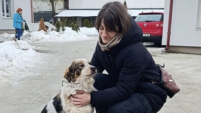 Жінка на вулиці присіла поруч із собакою Плюшкою з ЛКП «Лев», яка щойно стала частиної її сім'ї. Плюшка спокійно дивиться на жінку, довірливо притулившись. Навколо засніжений двір