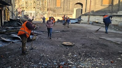 Прибирання пошкодженої території після обстрілу. На передньому плані розкидані уламки скла, цегли та металу. Кілька комунальників у помаранчевих жилетах зі світловідбивними смугами працюють із мітлами та лопатами, змітаючи дрібне сміття і згребаючи його в купи. На задньому плані — будівлі із забитими вікнами, складені купи металевих конструкцій, уламки покрівель.