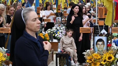 Люди, серед яких монахиня на передньому плані та молода жінка з дитиною, зібралися на Личаківському кладовищі серед військових поховань з українськими прапорами, хрестами та квітами. Багато присутніх тримають руки на серці під час молитви.