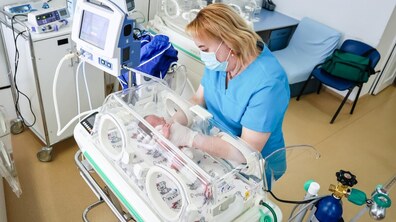 Медична працівниця у халаті й масці доглядає за новонародженим немовлям у відділенні інтенсивної терапії. Дитина лежить у прозорому кувезі, під’єднаному до медичного обладнання. Навколо є апаратура для моніторингу стану і підтримки життєвих функцій.