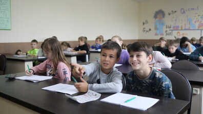 Учні сидять за партами під час уроку. За першою партою троє учнів. Дівчина пише щось на листочку. Один хлопчик вказує пальцем іншому на щось попереду. Діти усміхаються