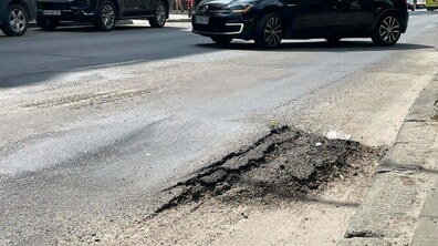 Вулиця Зелена. На передньому плані видно вибоїну та зруйноване дорожнє покриття — асфальт потріскався, частина поверхні просіла. На задньому плані рухаються автомобілі, поруч розташовані будинки та тротуар.