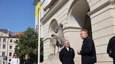 Біля входу до львівської ратуші міський голова Андрій Садовий спілкується з посолкою Канади в Україні Наталкою Цмоць на тлі аркадної стіни будівлі, кам’яної скульптури лева та піднятого українського прапора. Поруч проходять люди. Погода - сонячна.