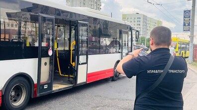 Інспектор у чорній футболці з написом «Управління транспорту» фотографує на смартфон міський автобус із відкритими дверима. Транспортний засіб припаркований біля тротуару на тлі міських багатоповерхівок.