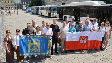 На площі Ринок стоять учасники делегації з естонського міста Тарту і представники Львівської міської ради та тримають прапори обох міст. Позаду них видно 4 автобуси