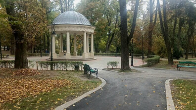 Ротонда у міському парку. До неї веде нова асфальтована доріжка, обабіч якої розміщені лавки. На землі опале жовте листя. Довкола високі дерева.