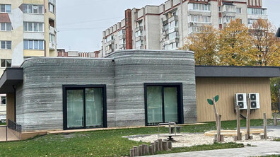 Одноповерхова будівля першої в Україні 3D-школи у Львові на тлі багатоповерхових житлових будинків. Перед будівлею пішохідні доріжки, газон і дитячий майданчик з ігровими елементами