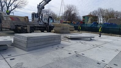 будівельний майданчик біля моста на вул. Ковча: кран піднімає бетонні плити. На землі — рівно викладені залізобетонні плити з отворами. Поруч кілька робітників у сигнальних жилетах. На фоні — паркан, дерева без листя й невеликі будівлі.