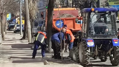 Прибирання міської вулиці у сонячний день. Біля тротуару стоїть помаранчевий самоскид, поруч невеликий синій трактор, а кілька комунальників у робочому одязі з лопатами та мітлами згрібають пісок і бруд уздовж бордюру. Частину змету вже зібрали в купу біля дороги, звідки його завантажують у вантажівку. На задньому плані видно дерева без листя, житлові будинки та припарковані автомобілі.