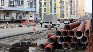 Вулиця Залізнична, де тривають ремонтні роботи. На передньому плані складені великі помаранчеві пластикові труби для прокладання комунікацій, поруч лежать бетонні плити та будівельні матеріали. Уздовж дороги встановлені тимчасові дорожні стовпчики з червоно-білими смугами, а поряд видно розриту ділянку землі. На дорозі стоїть невелика вантажівка, поруч ідуть кілька людей, один із них у світловідбивному жилеті. На задньому плані — сучасні багатоквартирні будинки з яскравими фасадами різних кольорів.