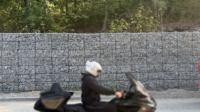 Підпірна стінка з прямокутних металевих сіток, щільно заповнених сірим колотим каменем (габіонів). Попереду стіни вздовж асфальтованої дороги на передньому плані проїжджає великий чорний мотоцикліст у темному костюмі та білому шоломі.