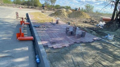 Процес укладання тротуару на вул. Бескидській. Прямокутна ділянка, частково викладена рожевою плиткою. Край плитки не завершений, перед ним — підготовлений шар сірого піску. На задньому плані купи піску, два екскаватори та зелені дерева.