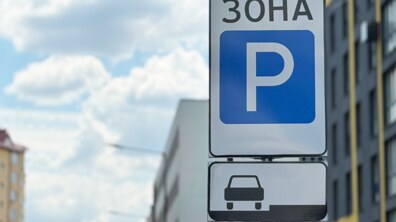 Крупним планом зображено дорожній знак паркувальної зони. На білому прямокутному щиті написано «ЗОНА», а нижче розміщений великий синій квадрат із білою літерою «P», що позначає місце для паркування. Під основним знаком встановлена додаткова табличка із силуетом легкового автомобіля, який припаркований паралельно бордюру.