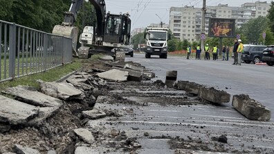 Ремонтні роботи на вул. Мазепи. На передньому плані видно розбитий асфальт і демонтовані бетонні плити вздовж узбіччя. Екскаватор знімає старе покриття біля металевої огорожі, поруч стоїть вантажівка. Далі на дорозі стоять кілька працівників у світловідбивних жилетах. На тлі — багатоповерхівки, електроопори та рекламний білборд.