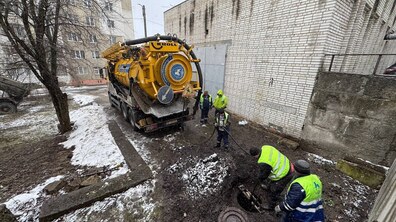 Процес промивки каналізації в зимових умовах. У центрі кадру — велика жовта вакуумна машина марки Kroll. На задній частині цистерни видно великий круглий люк, намотані шланги та дорожній знак. П'ятеро працівників у спецодязі зосереджені навколо відкритого каналізаційного люка: двоє робітників нахилилися безпосередньо над відкритим колодязем, а ще троє стоять поруч, контролюючи процес подачі шланга від машини в люк.