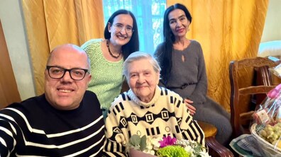 Ювілярка, її родичі та працівник управління соціального захисту усміхаються, позуючи для спільного фото. Пані Ніна тримає в руках букет квітів, а поруч видно кошик з фруктами.