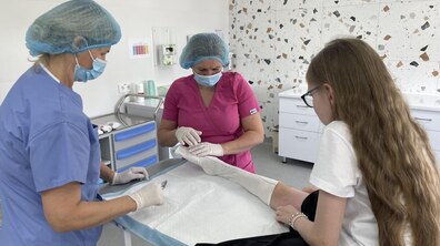 Медична процедура у лікарняному кабінеті. Дві медичні працівниці в захисному одязі, масках, шапочках і рукавичках оглядають ногу 11-річної пацієнтки, яка сидить спиною до камери. Видно, що на нозі є білі еластичні бинти.