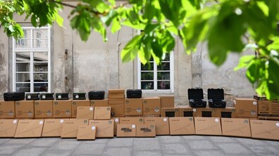 Уздовж стіни акуратно складено багато коробок із написом “Starlink” та технічне обладнання. Верх кадру закриває зелене листя дерева, крізь яке пробивається сонце.