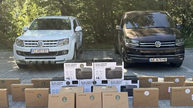 На відкритому майданчику стоять два автомобілі, а перед ними – багато коробок зі Starlink та портативними зарядними станціями, що підготували для передачі військовим. Позаду видно зелені дерева.