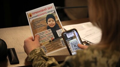 Людина у військовій формі тримає листівку з інформацією про терміновий збір коштів на лікування маленької дитини. Поруч вона сканує QR-код зі смартфона, щоб долучитися до допомоги.