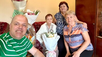 Ювілярка Ніна Калиновська, дві її родички та працівник управління соціального захисту Іван Фасольник сидять у кімнаті: ювілярка тримає в руках великий букет квітів, а позаду на столі стоять ще два букети та «Кошик довголіття» зі смаколиками. Усі радісні й усміхнені.