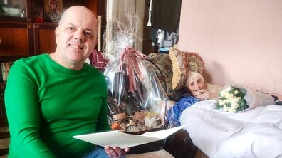 Столітня ювілярка Ніна Телешевська лежить на ліжку в кімнаті, біля неї сидить працівник Іван Фасольник, який тримає в руках вітальний лист та кошик з фруктами