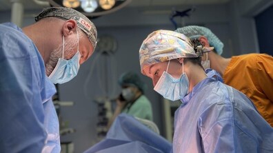 Команда медичних працівників оперує в операційній. Двоє хірургів у стерильному одязі, масках і медичних шапочках зосереджено працюють біля операційного столу. На задньому плані видно інших медиків та хірургічне обладнання.