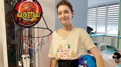 Усміхнена 14-річна Настя стоїть біля баскетбольного кошика, що у реабілітаційному залі лікарні; дівчинка одягнена у футболку світлого кольору, під рукою тримає м’яч